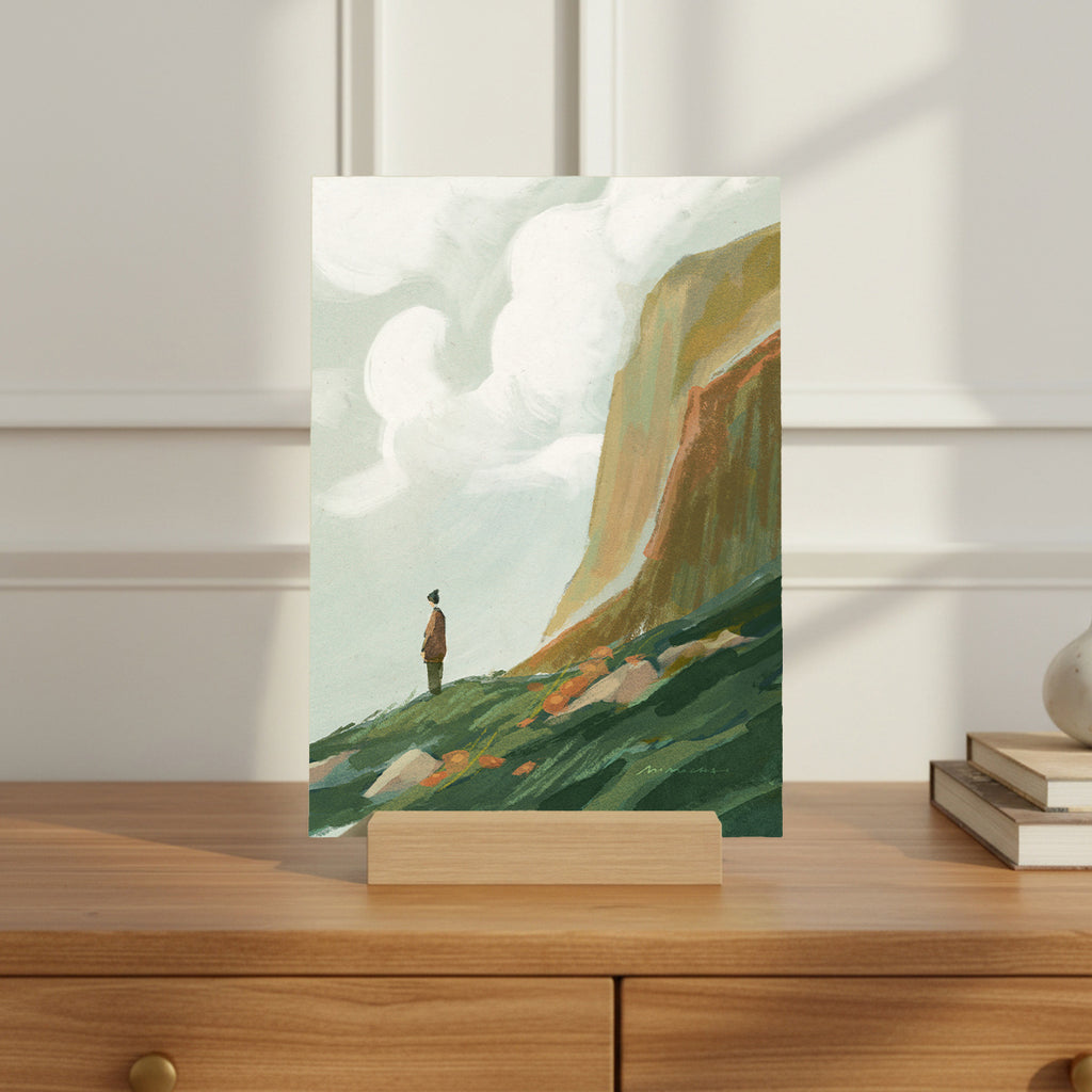 Art print of a person standing on a grassy hill with a mountain in the background, displayed on a wooden stand.