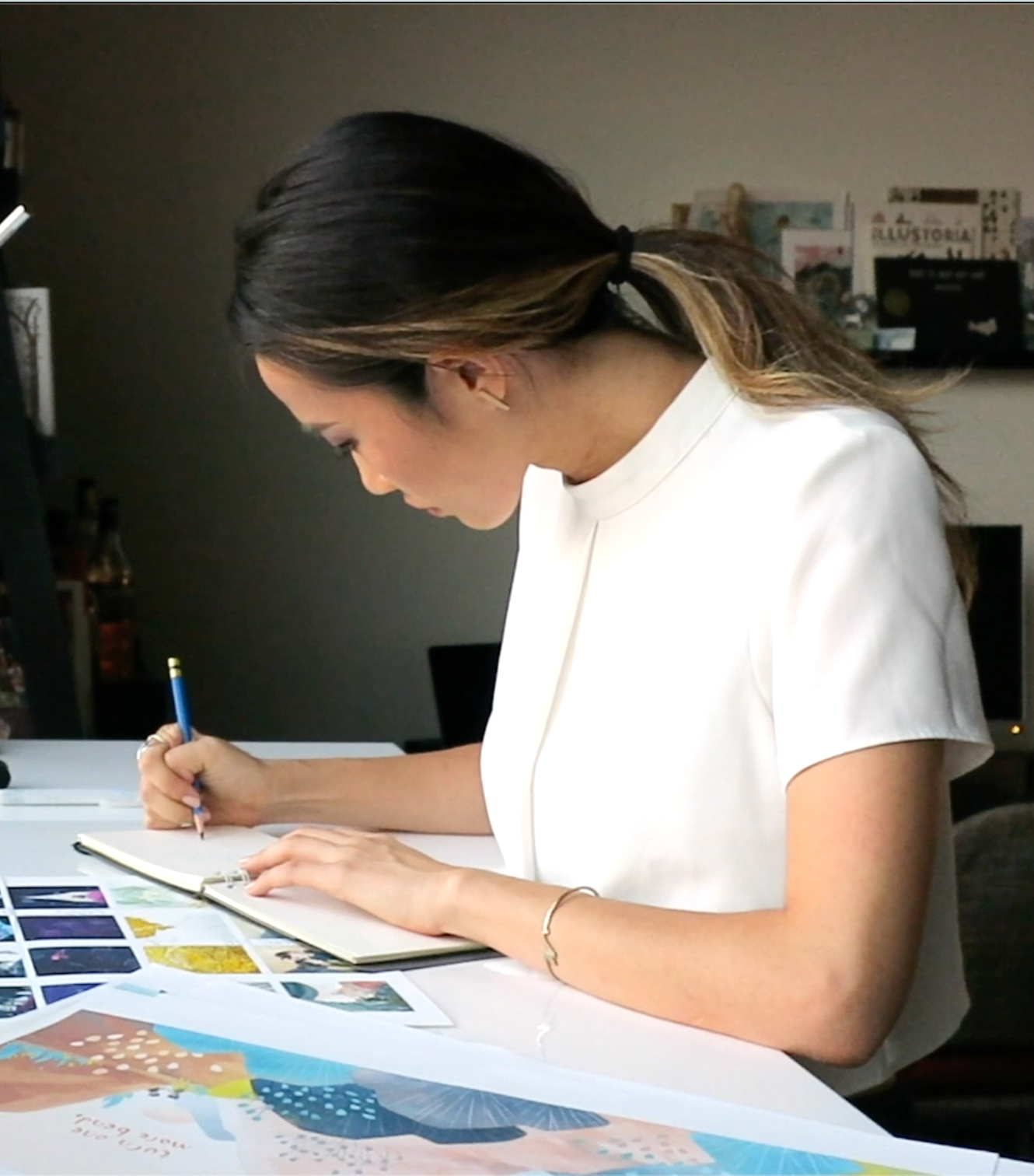 A side view of a woman drawing in a sketchbook at a table with illustrations around her