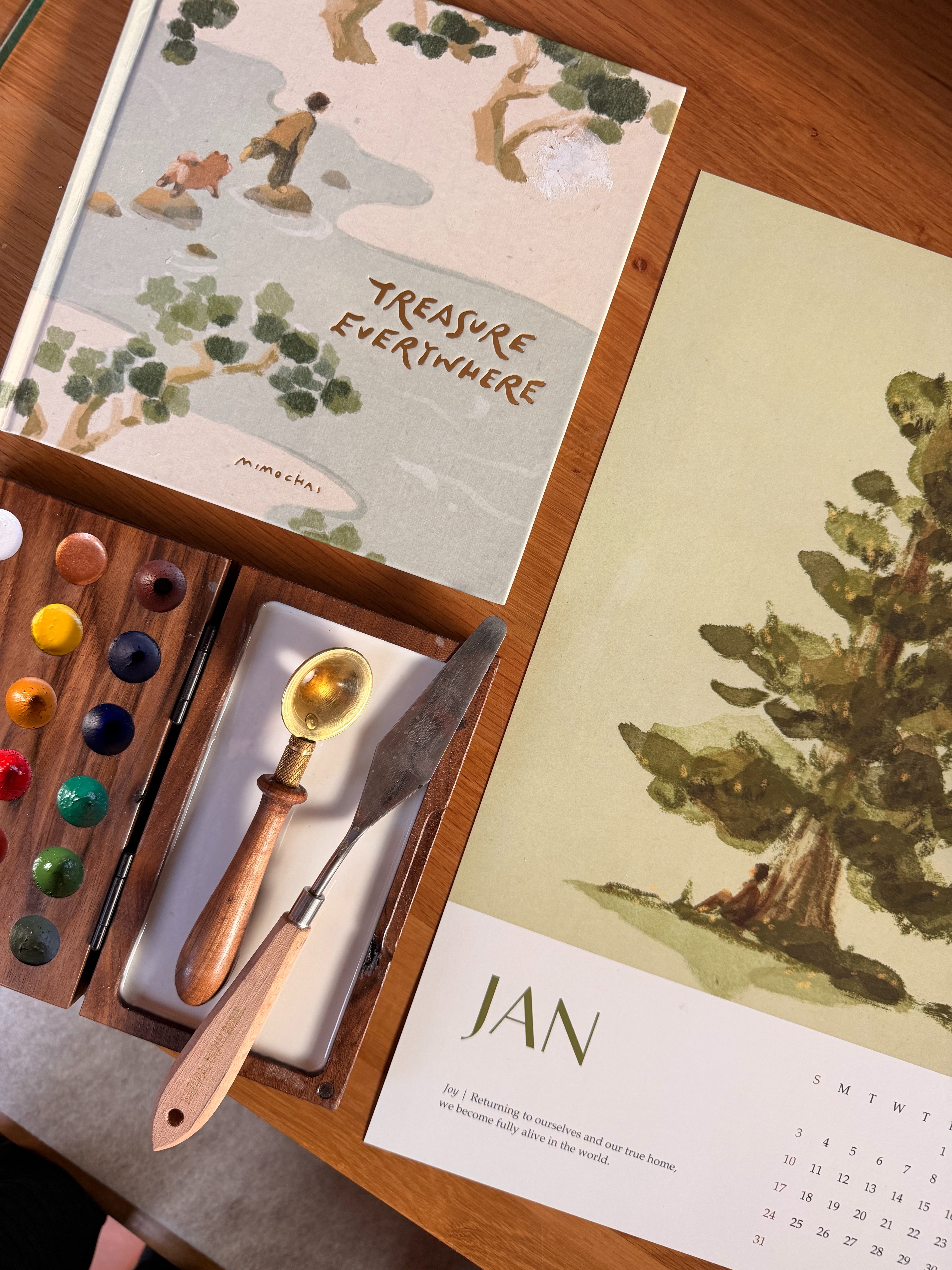 Art supplies including a paint palette, brushes, and a calendar on a wooden surface.