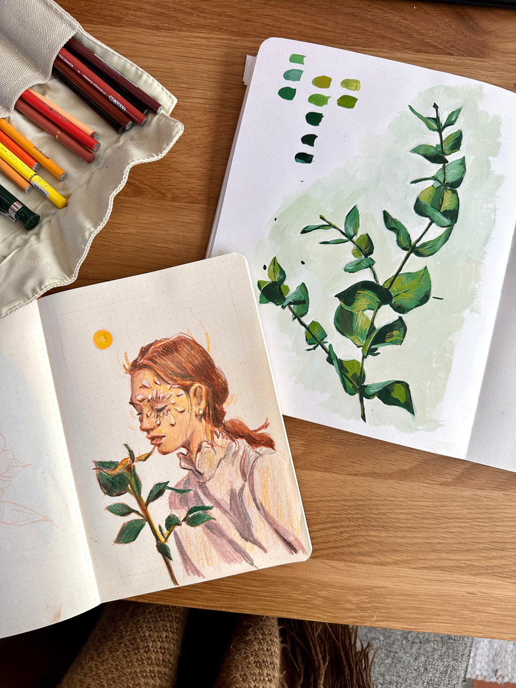 Sketchbooks on a desk showing colored pencil and gouache drawings of a woman and plants